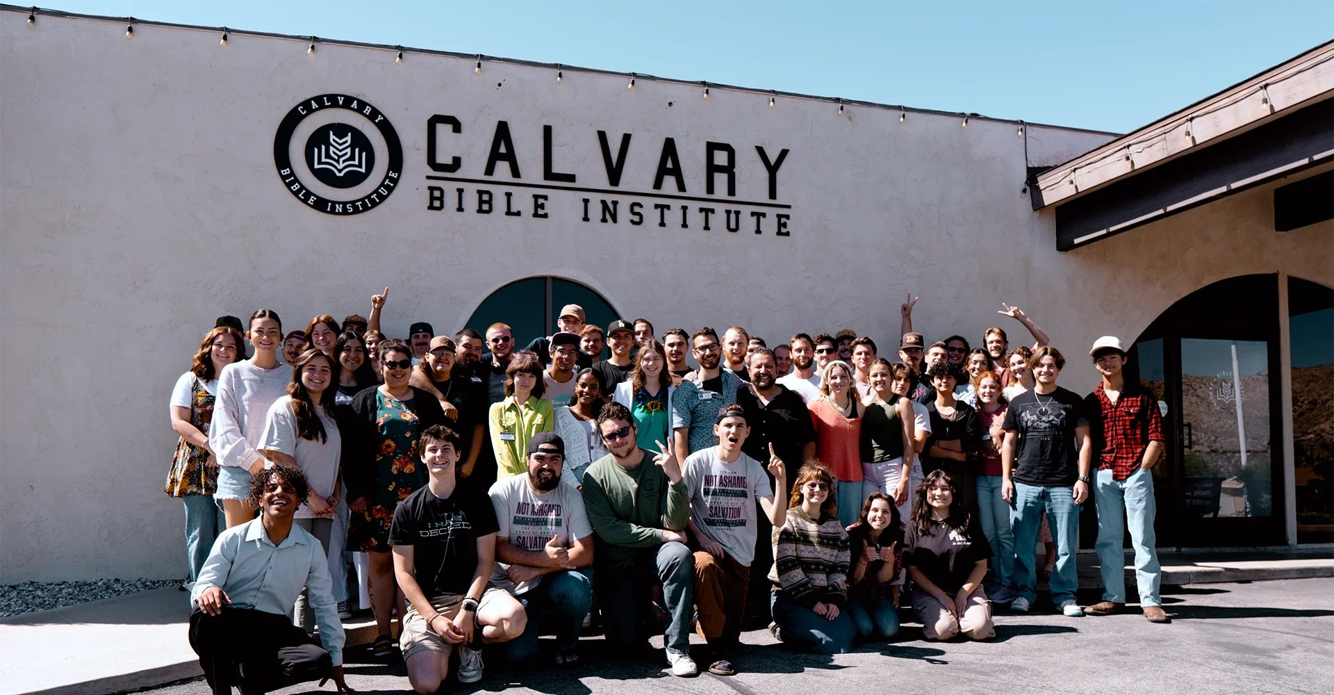 A large group of diverse people standing in front of the Calvary Bible Institute building. The group is smiling and posing for a photo on a sunny day.