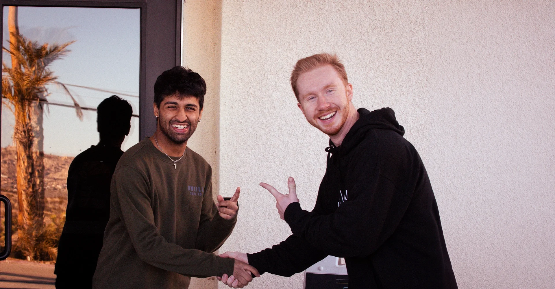 Two young men shaking hands and smiling indoors, with a window showing a desert landscape with palm trees in the background.