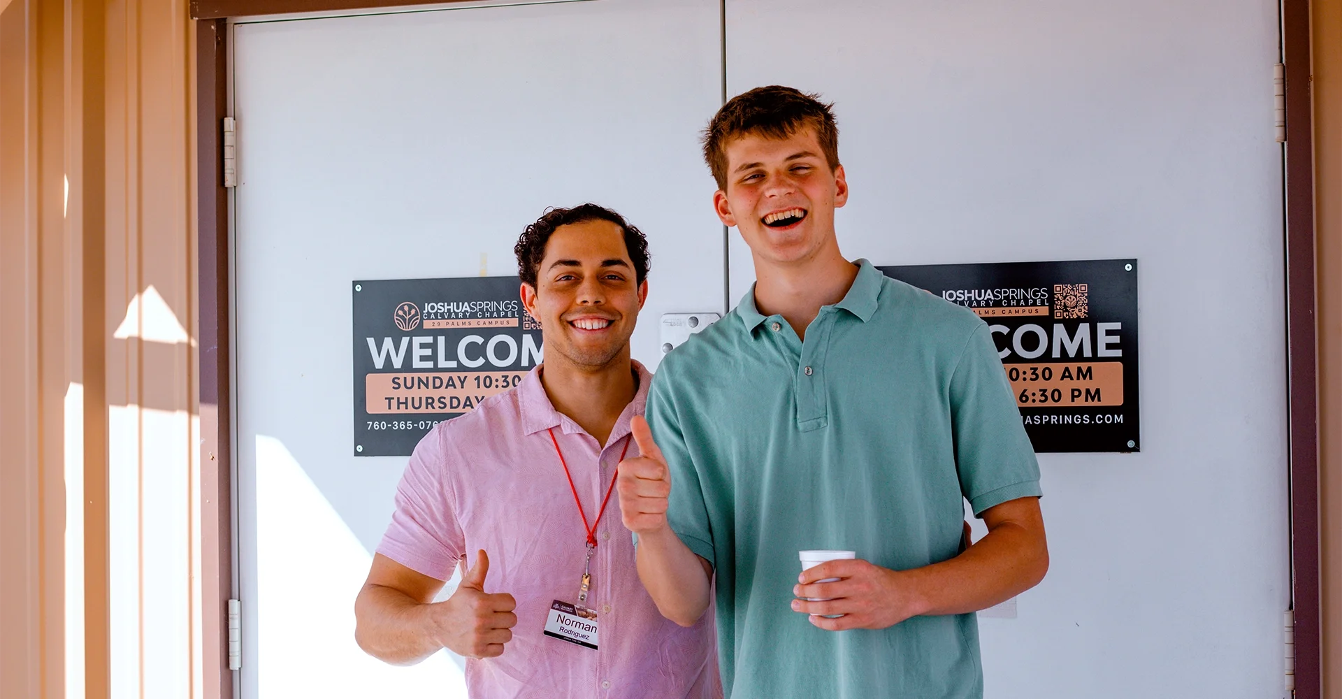 Two smiling men standing in front of a door with welcome signs for Joshua Springs Calvary Chapel, one wearing a pink shirt with a name tag, and the other wearing a teal polo shirt holding a cup.
