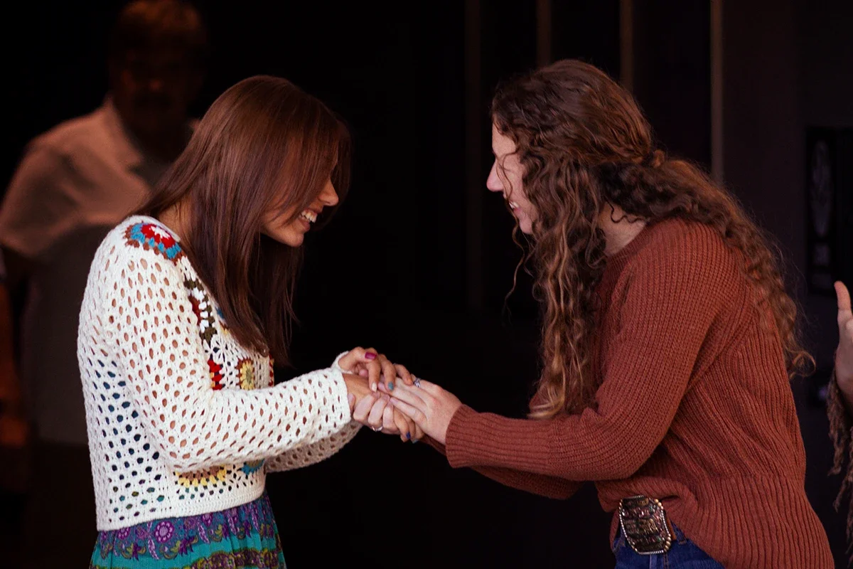 Two women are smiling and holding hands in a warm, joyful exchange. One woman has brown hair and is wearing a white crocheted sweater with colorful patterns, while the other has curly hair and is dressed in a brown sweater. They appear to be in an indoor setting with a dark background.