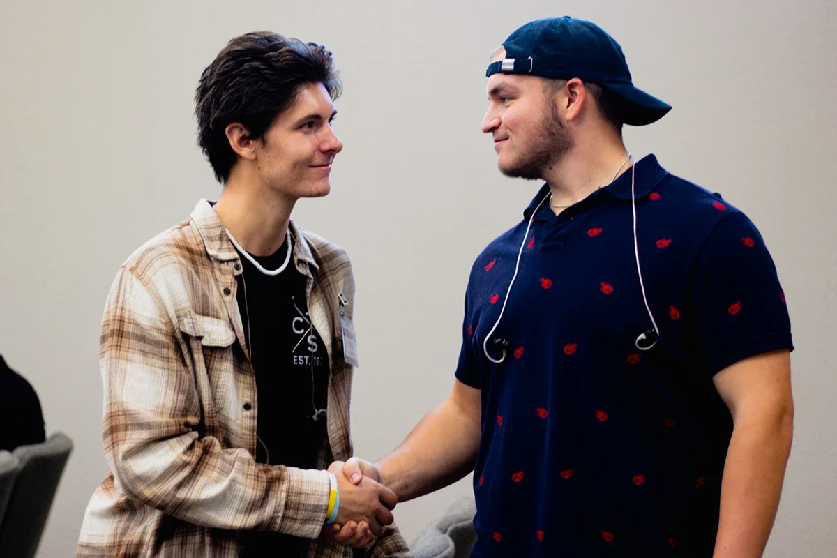 Two young men shaking hands and looking at each other. One is wearing a beige plaid shirt over a black t-shirt, and the other is wearing a navy blue polo with red designs and a backwards baseball cap. They both have earphones around their necks.