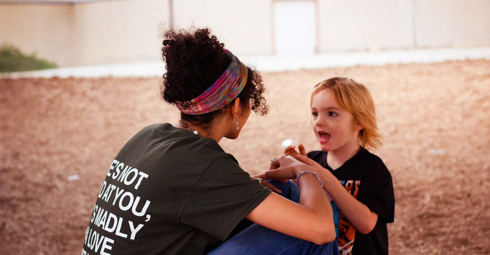 A woman with curly hair, wearing a colorful headband and a black T-shirt, is holding hands with a young boy with red hair and a black T-shirt, having an animated conversation inside a gymnasium.
