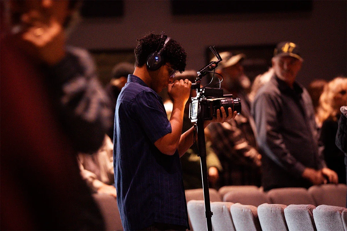 A young man wearing headphones and glasses operating a professional video camera at an indoor event surrounded by seated and standing attendees.