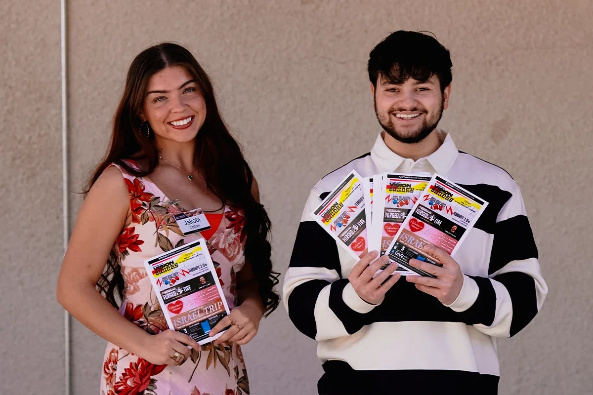 Two smiling people standing side by side holding flyers for a conference or event, with a beige wall in the background.