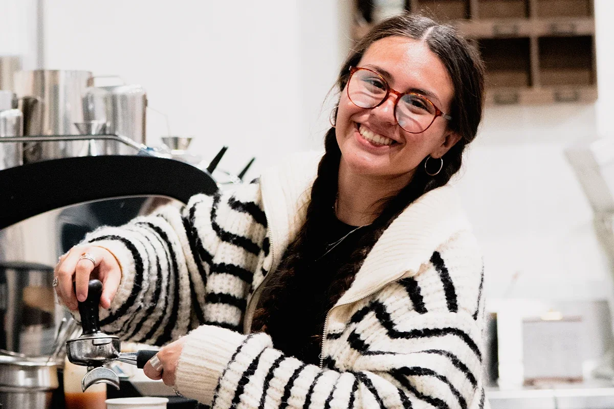 A smiling woman with glasses and long dark hair braided, wearing a black and white striped sweater, preparing a coffee in a kitchen.