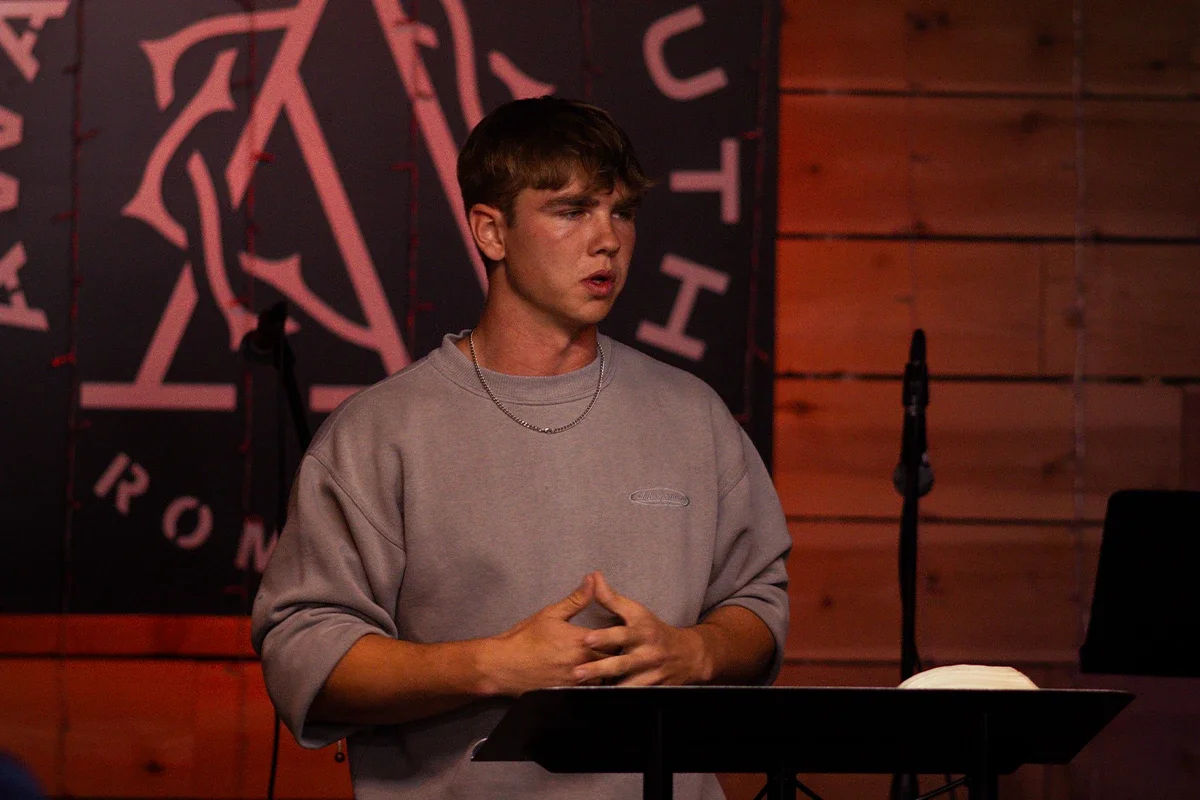 A young man standing at a music stand, speaking in front of a microphone in a dimly lit room with wood paneling. Behind him is a black banner with pink lettering and a graphic design.