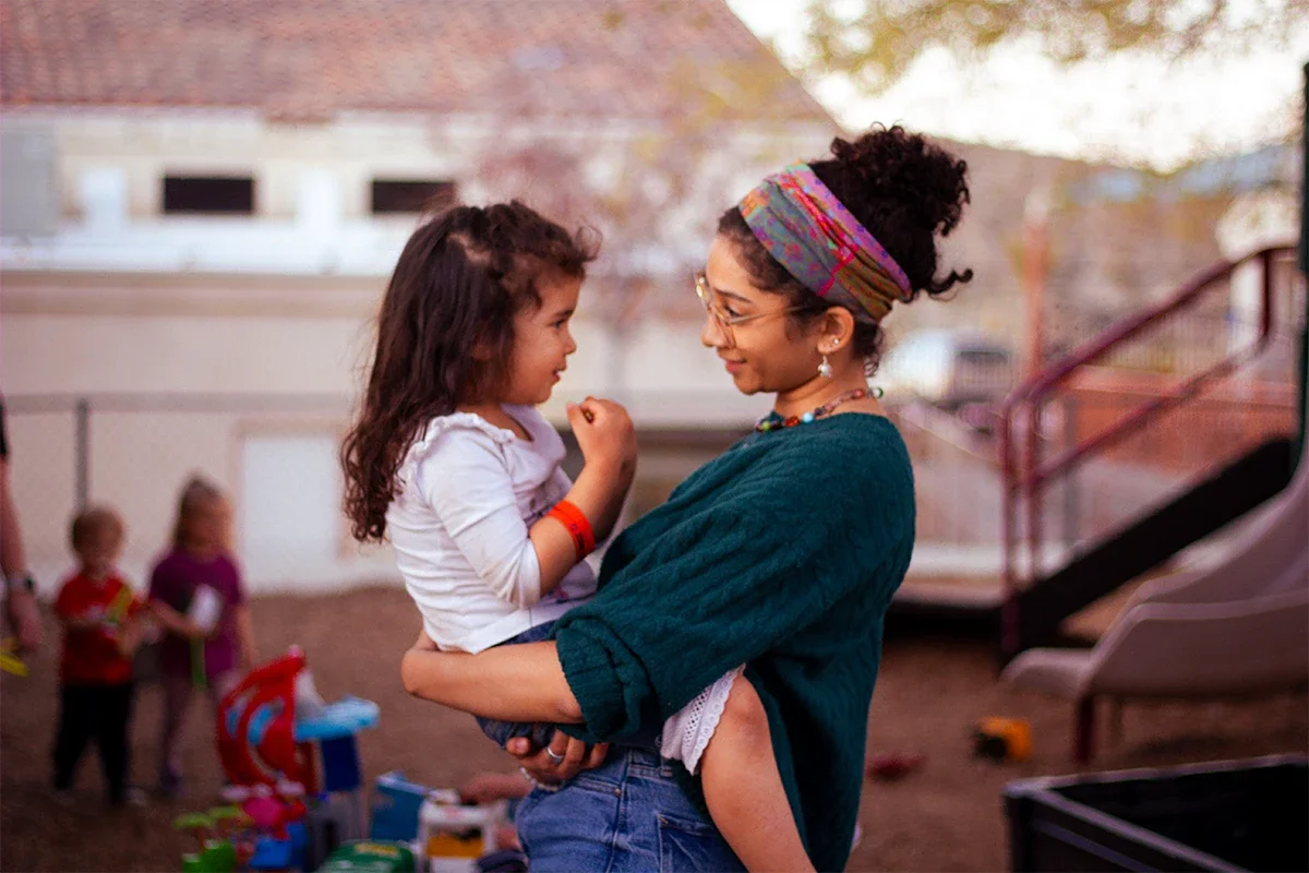 A woman with glasses and a colorful headband holding a young girl in her arms, smiling and looking at each other outdoors during what appears to be early evening. There are children playing in the background.