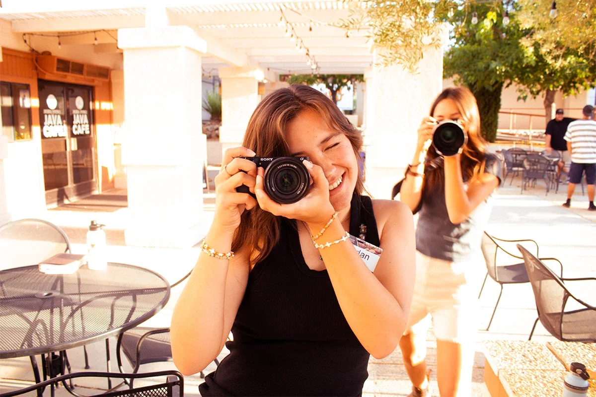 A young woman holding a camera up to her face and smiling, with another woman in the background taking a photo.