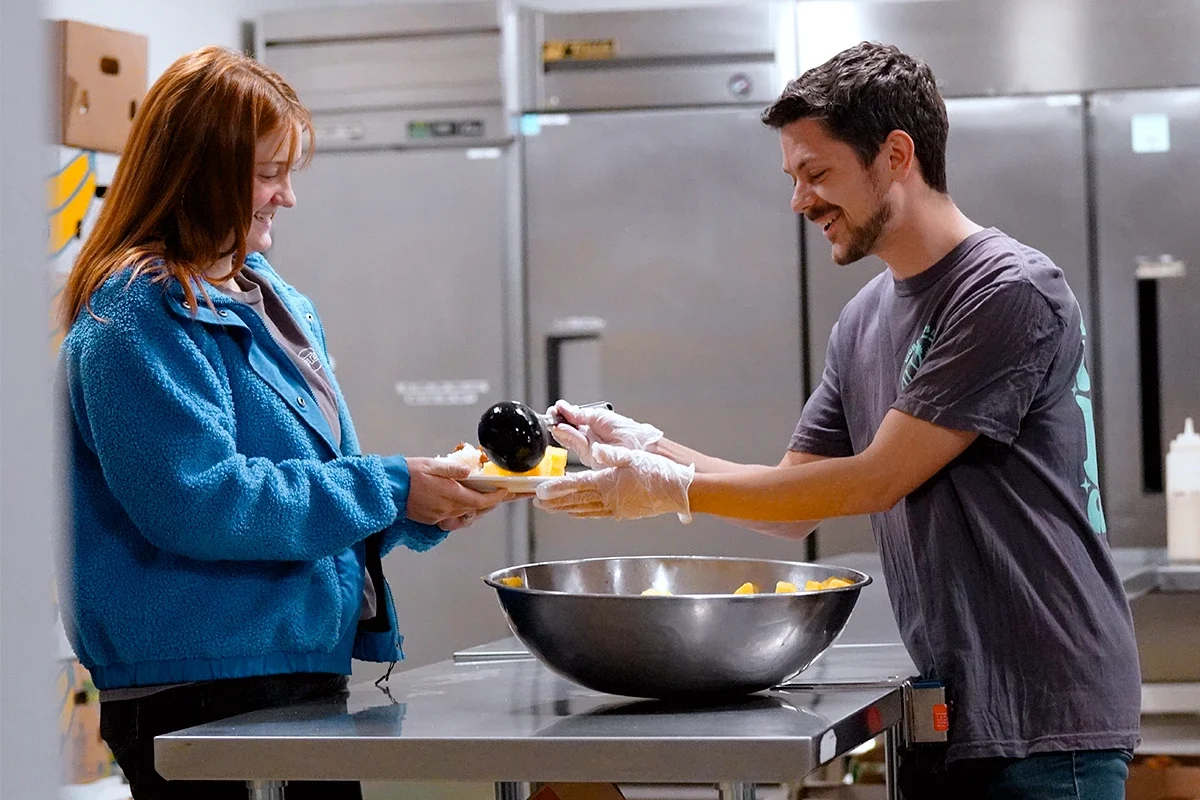 A man with short dark hair and a beard is handing a plate of food to a woman with long red hair in a commercial kitchen. The man is wearing a gray t-shirt and transparent gloves, while the woman is wearing a blue fleece jacket. There is a large stainless steel bowl on the table in front of them, and industrial refrigerators in the background.