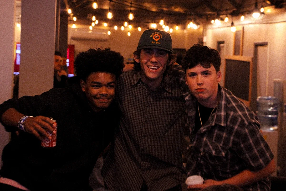 Three young men posing together in a dimly lit room with string lights hanging from the ceiling; they are smiling and appear to be at a social gathering or party.