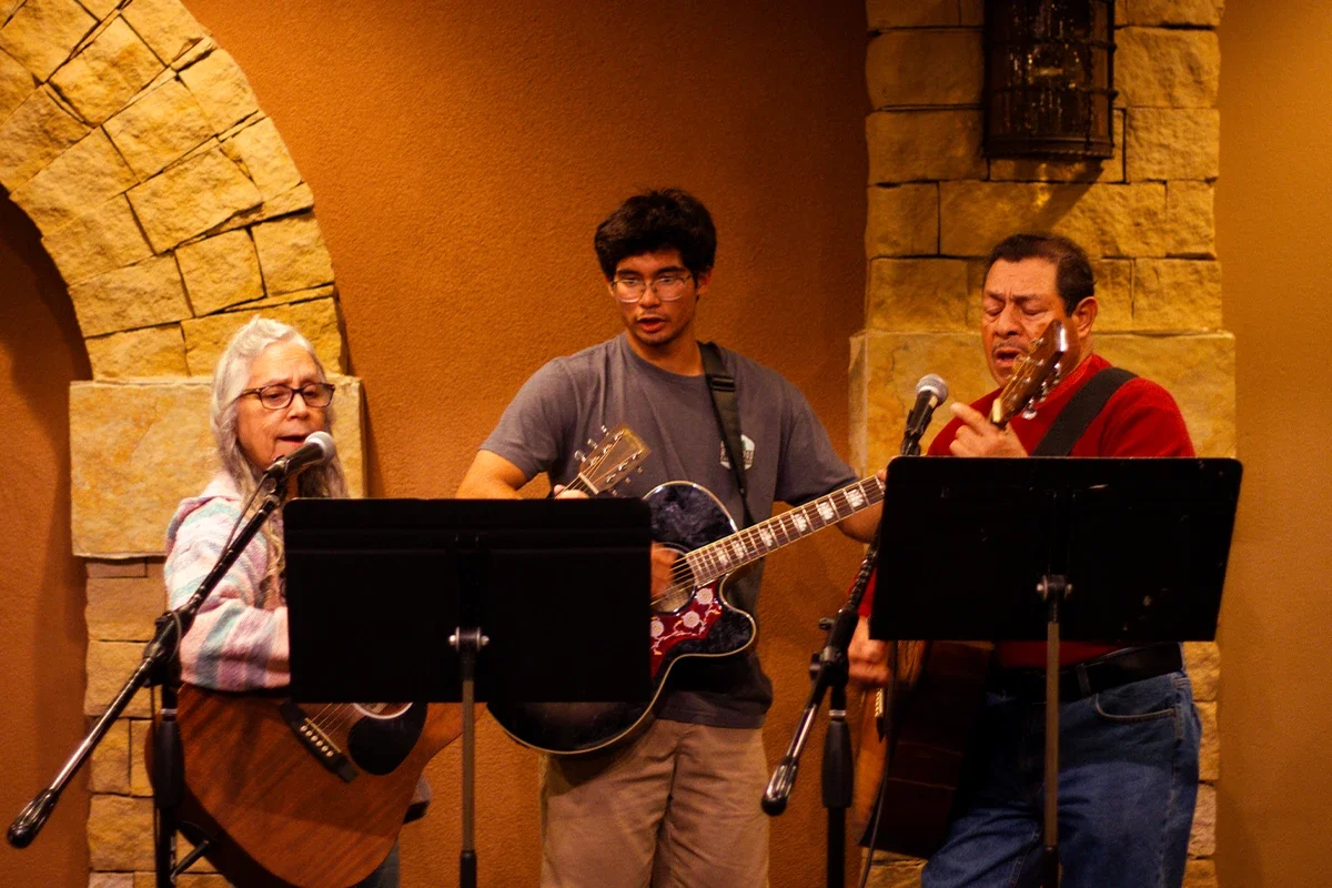Three people singing and playing guitars on stage with microphones and music stands in front of them.