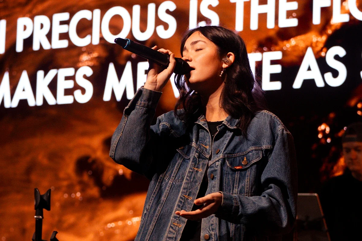 A woman with dark wavy hair singing into a microphone on stage, wearing a denim jacket. Behind her is a large screen with white text on a fiery orange background.