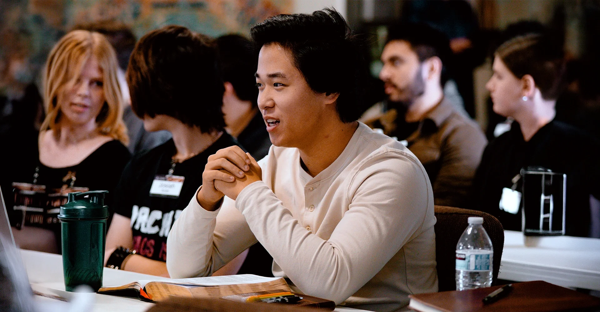 A young man with black hair sitting at a table during a conference or seminar, speaking or asking a question, with other attendees around him.