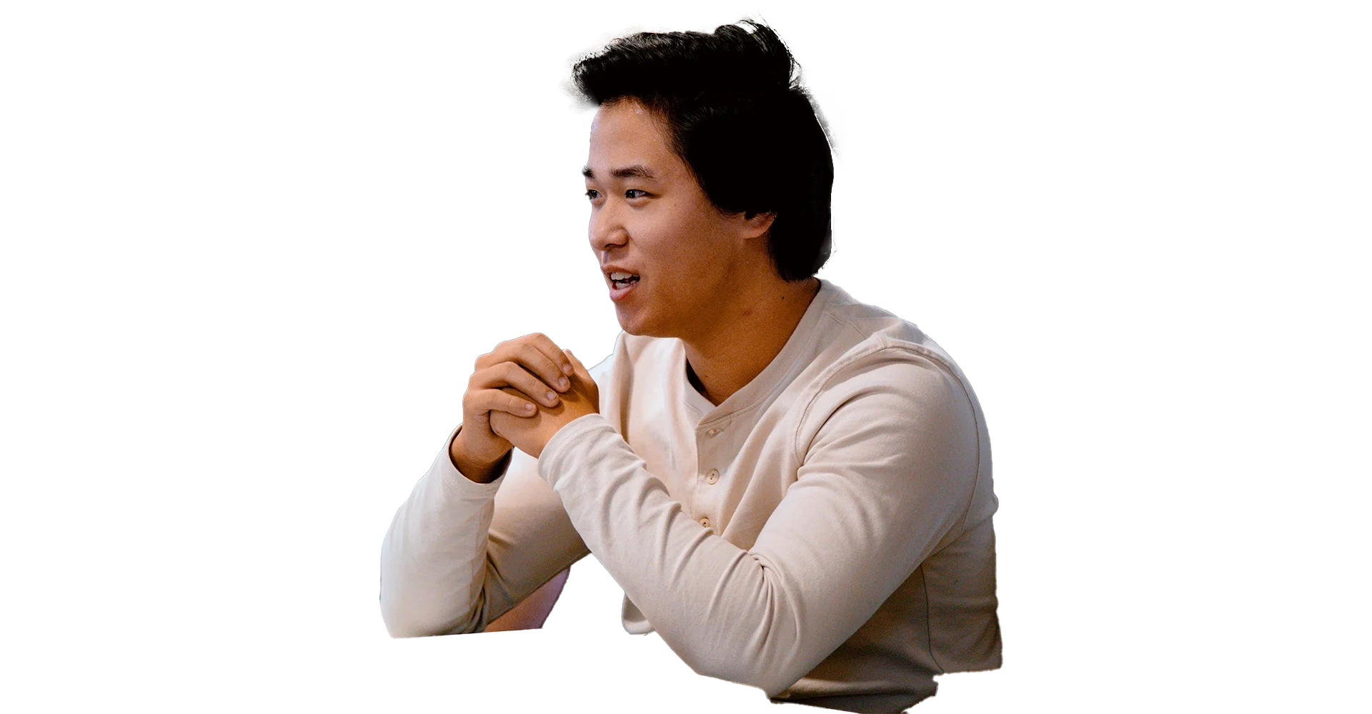 Young man with short black hair sitting at a table, wearing a light-colored long-sleeve shirt, with hands clasped, smiling and looking to his right.