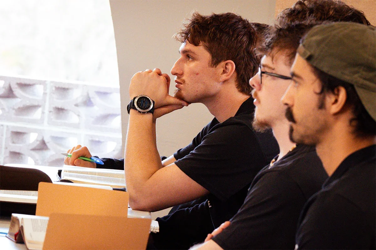 Three young men sitting at a desk in a classroom, attentively listening, with notebooks, and one wearing a baseball cap.