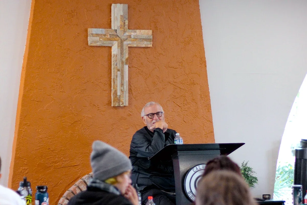 A man with glasses and gray hair sits at a black podium with a circular logo, in front of an orange wall with a wooden cross, attending a religious gathering or sermon.