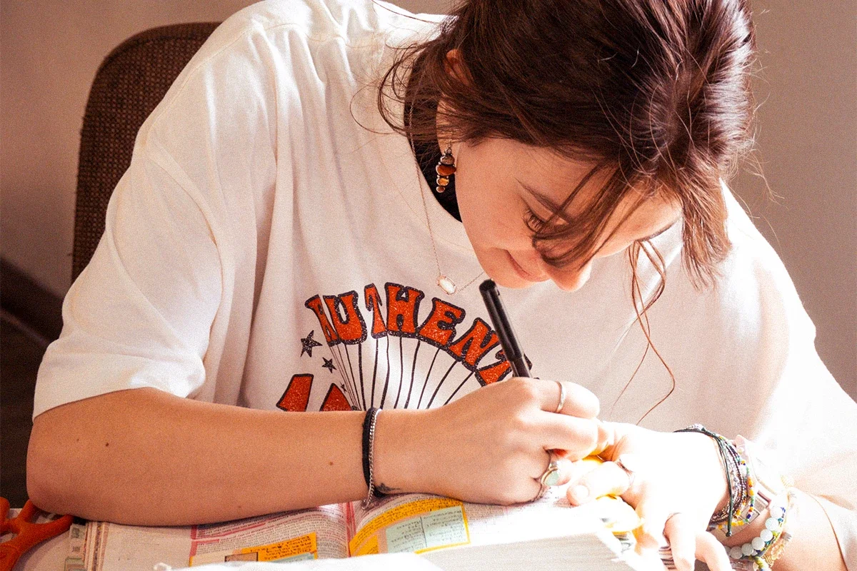 A young woman with short brown hair, wearing a white graphic T-shirt and multiple bracelets, is sitting at a desk, writing in a notebook with a black pen.