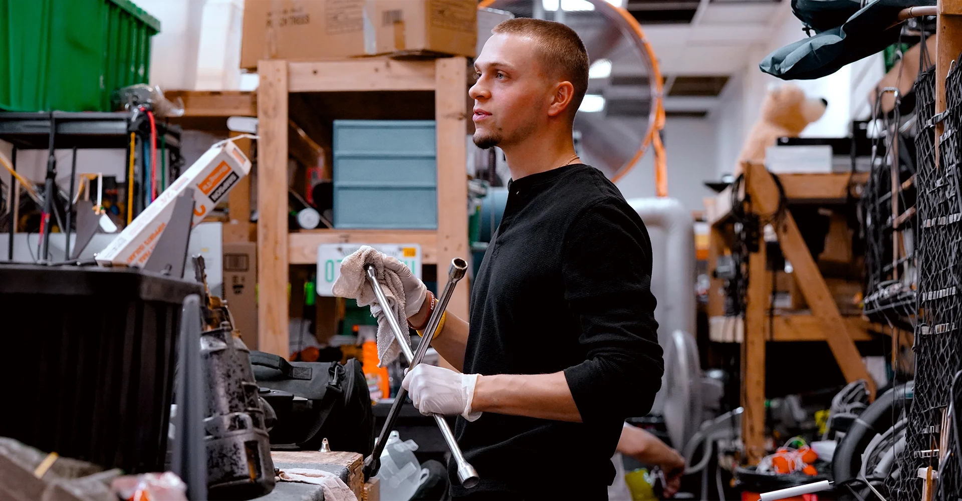 A young man with a buzz cut wearing a black long-sleeve shirt and white gloves is working in a cluttered workshop, holding a socket wrench and a cloth, with shelves of tools and boxes in the background.