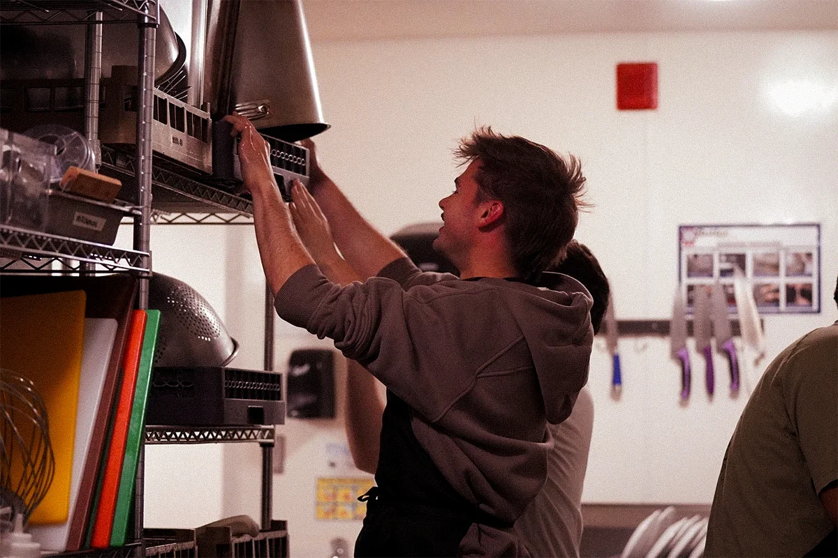 A young man reaches up to a high shelf in a kitchen or storage room, with various kitchen items and utensils visible on the shelves.