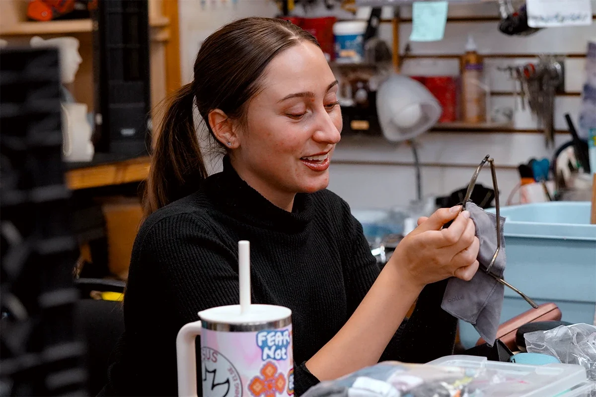 A woman with brown hair tied back in a ponytail, smiling and holding a pair of glasses in one hand and a gray cloth in the other. She is sitting at a cluttered workbench filled with various tools and supplies in a workshop or garage.