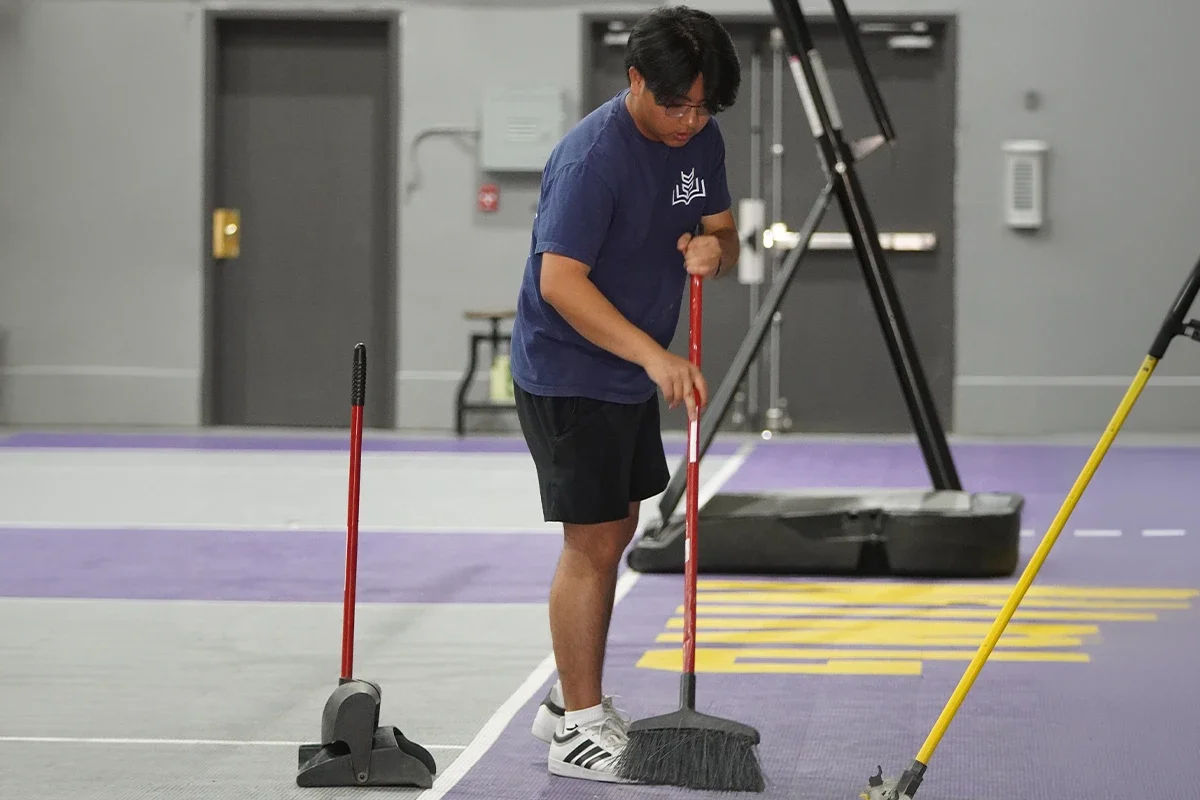 A person in a blue shirt, black shorts, and white Adidas sneakers sweeping the floor of an indoor sports court with a large push broom.