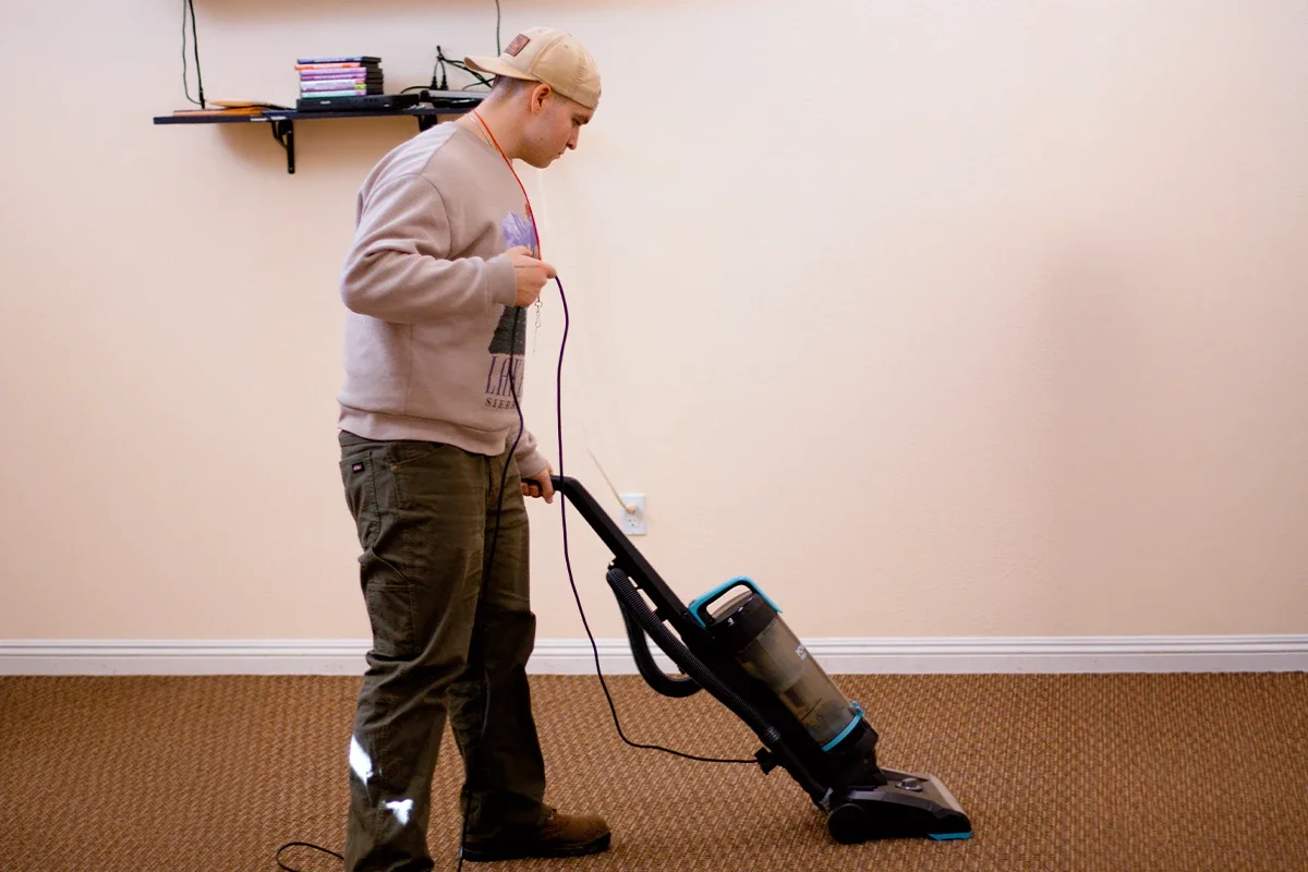 A man vacuuming a carpeted room with a black and blue vacuum cleaner, wearing a beige cap, beige sweatshirt, and dark pants.