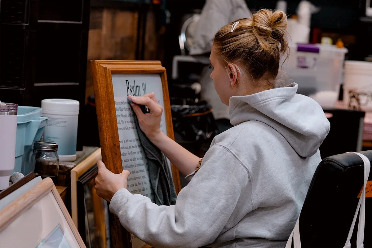 A woman with a bun hairstyle, wearing a light gray hoodie, writing on a framed whiteboard in a cluttered workspace.