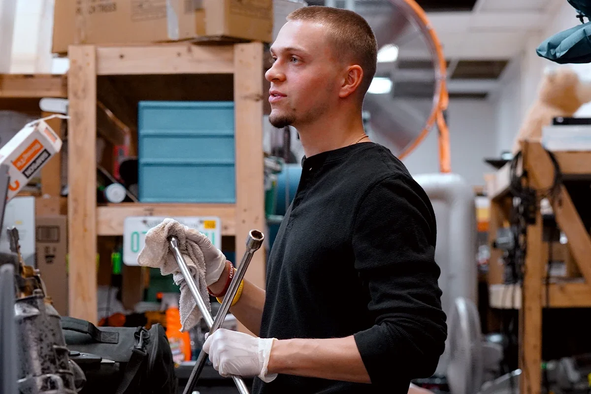 Young man cleaning a tool in workshop with wood shelves and storage boxes in background.