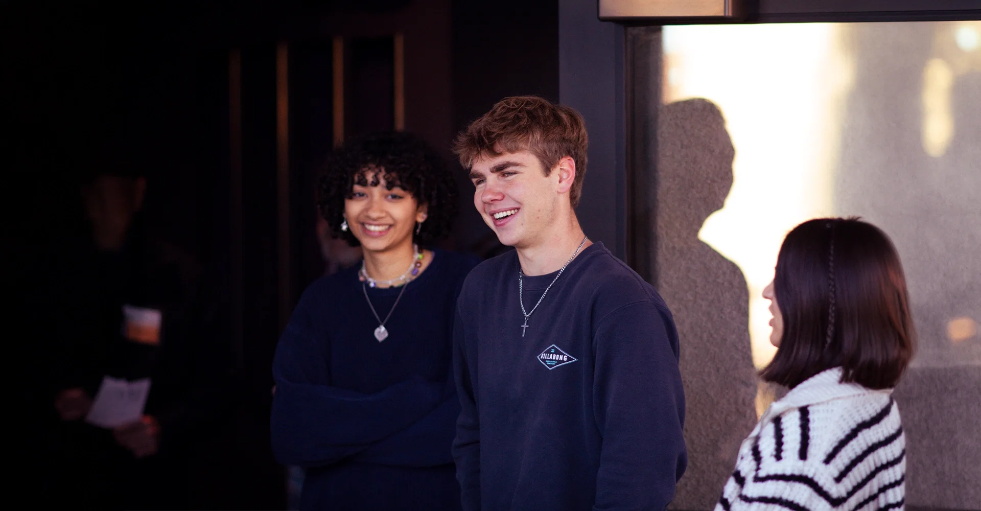 Three teenagers, two girls and one boy, smiling and chatting indoors. The boy wears a dark shirt with a cross necklace, and the girls have headphones and necklaces, with one wearing a striped sweater and the other wearing a dark top with layered necklaces. The background is softly lit, with a shadow of a girl on the wall.