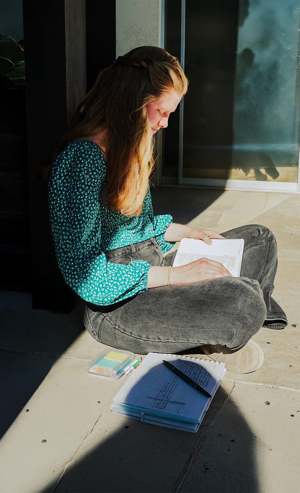 Young woman sitting on the ground reading a book in sunlight, with notebooks and pens nearby.