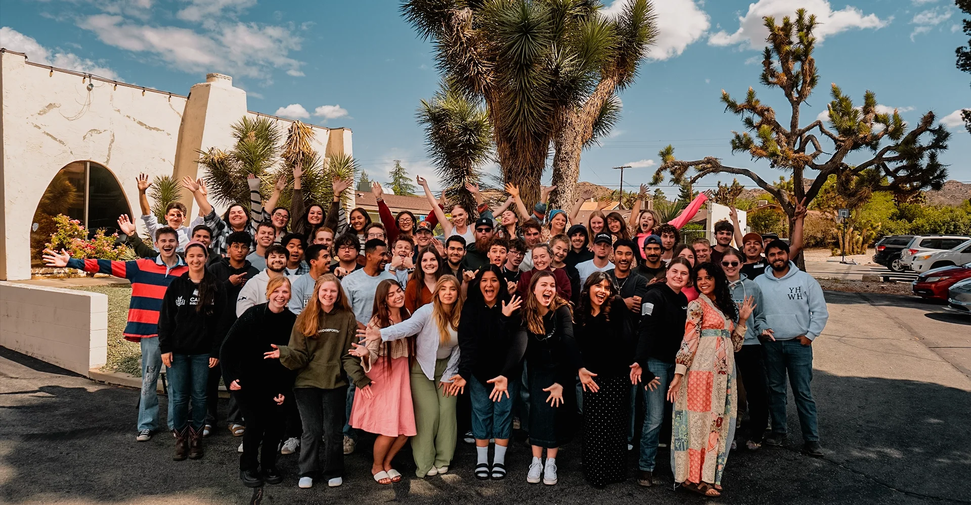 Group of diverse young people smiling and waving outdoors on a sunny day, with palm trees and parked cars in the background.