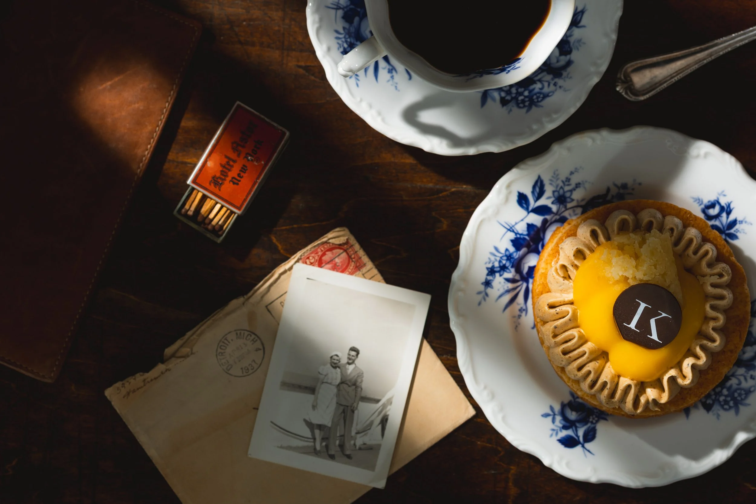 A vintage-style table setting with a cup of black coffee in a blue and white patterned cup and saucer, a plate with a Lemon Meringue tart, a small matchbox, an old envelope, and a black-and-white photograph of a couple posing on a boardwalk.