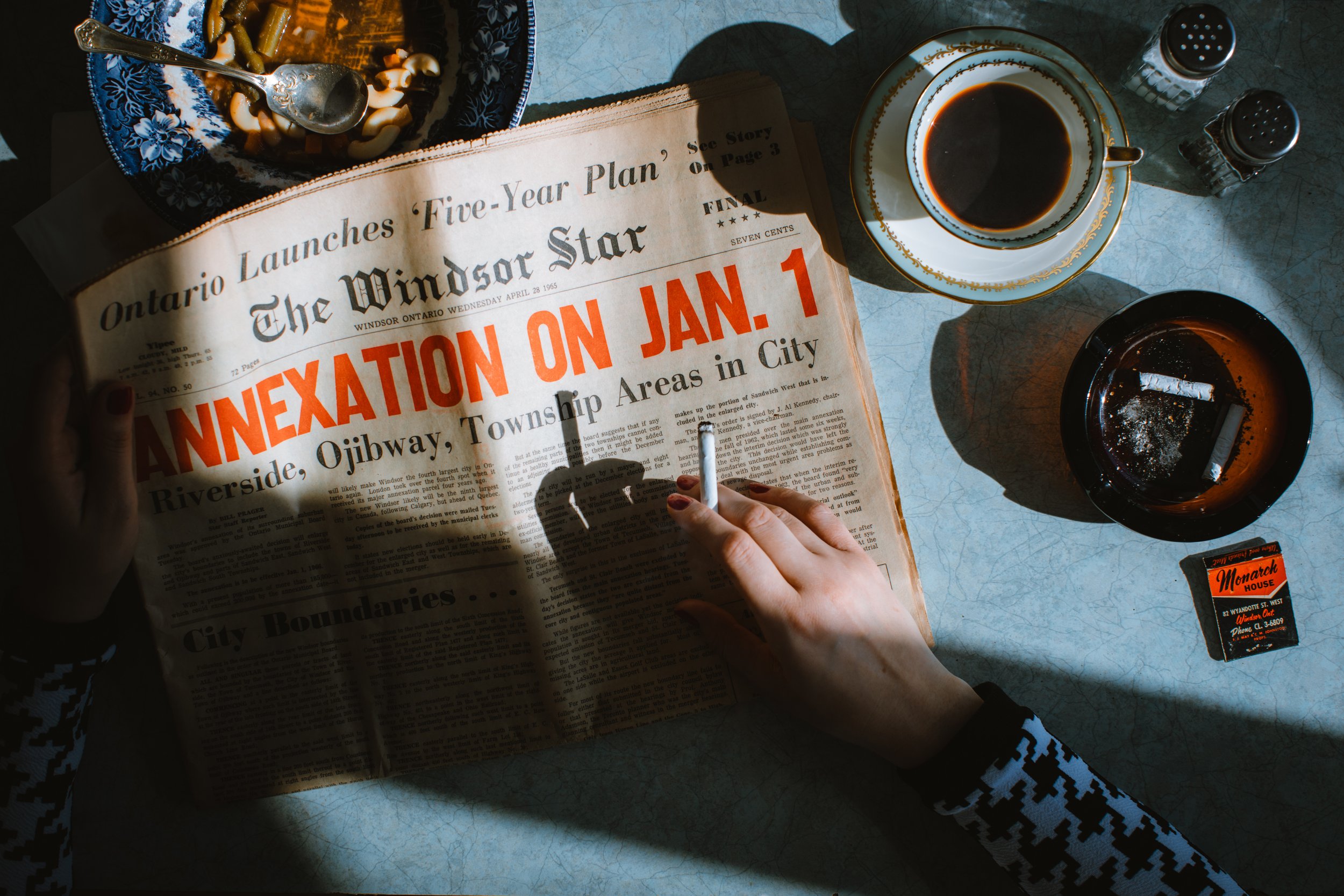 A person with painted red nails is reading a newspaper with a headline about annexation on January 1st, at a table with a cup of coffee, an ashtray with cigarettes, salt and pepper shakers, and a pack of Monarch house cigarettes.