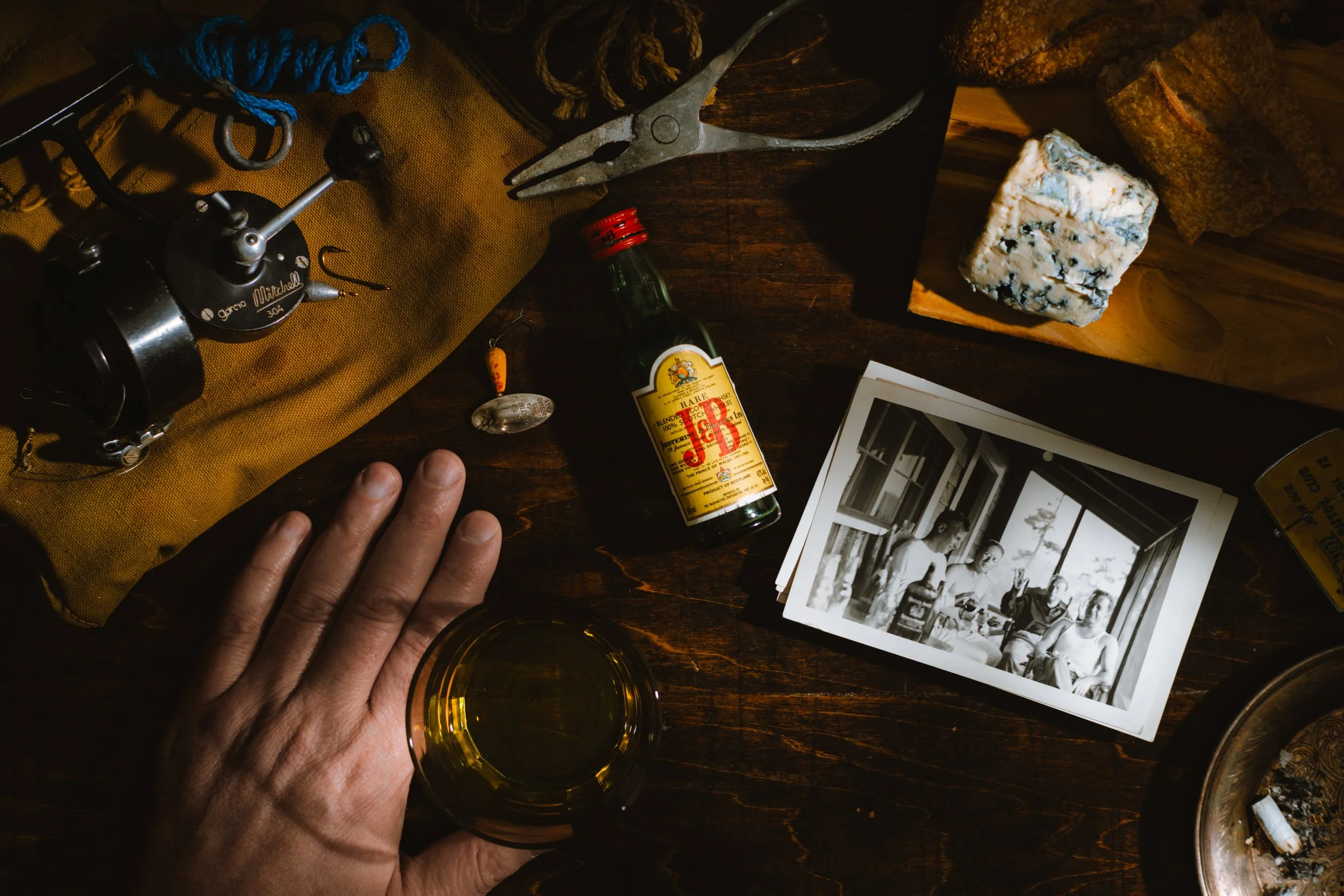 A wooden table with a black and white photograph of three people, a bottle of J&B whiskey, a glass of whiskey held by a hand, a vintage camera, a pair of pliers, a small container of cheese, cloths, and a few pieces of rope.