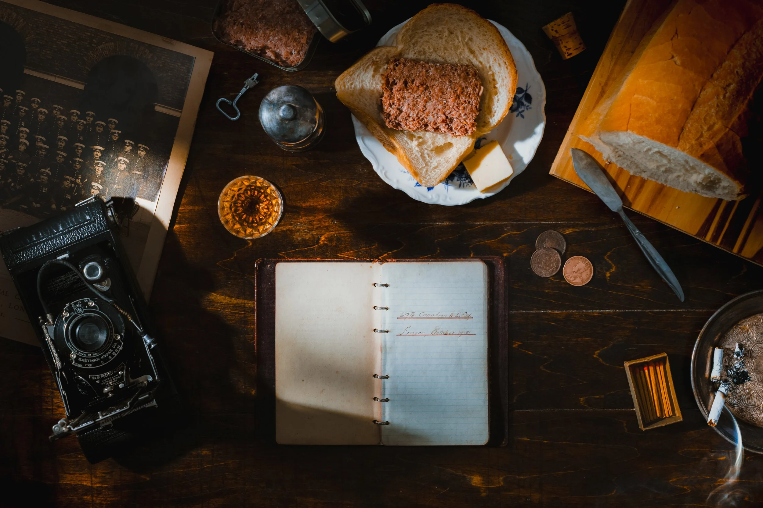 A vintage camera, a notebook with handwritten notes, a plate with sliced bread and a meatloaf, a glass of whiskey, some coins, a pack of cigarettes, a lighter, and a lit cigarette on an ashtray on a wooden table.