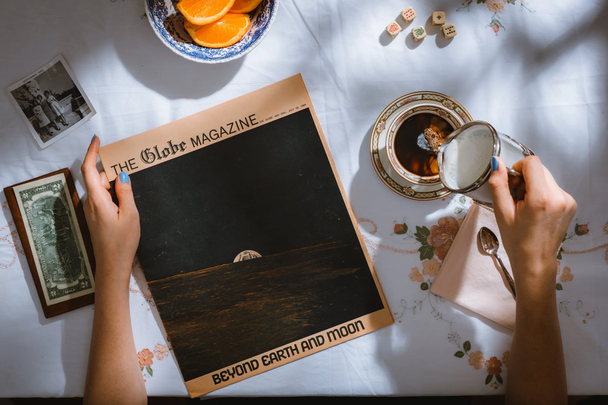 A person holding a black magazine titled 'Beyond Earth and Moon' on a white floral tablecloth, with a cup of coffee on a saucer, a glass of milk or cream, a bowl of oranges, a stack of dollar bills, a vintage black-and-white photograph, and a set of 