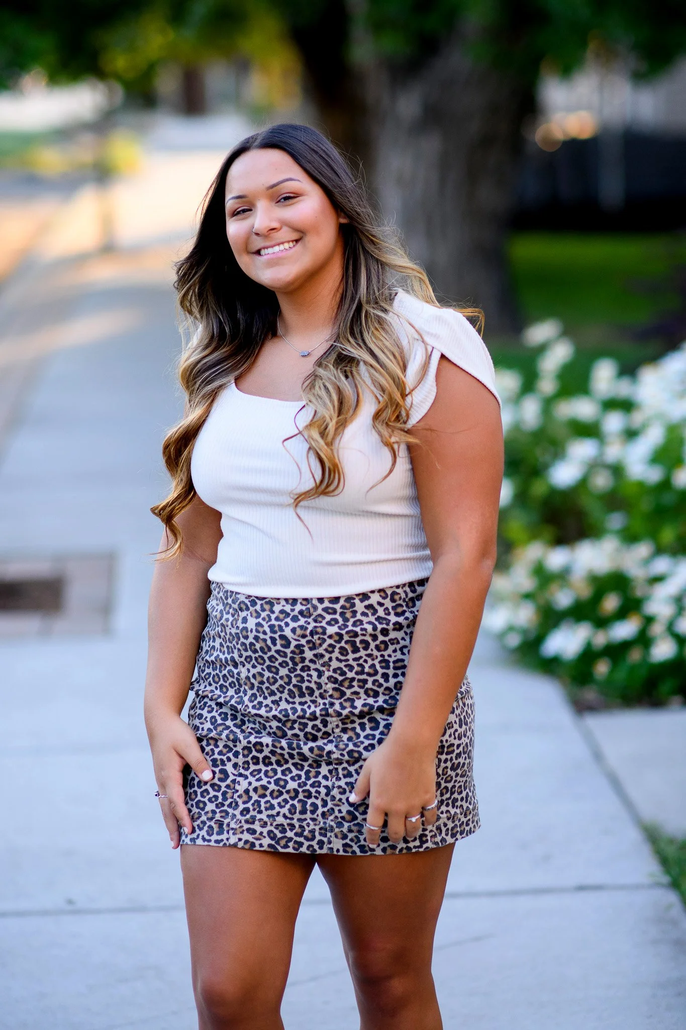 Young woman smiling outdoors with green trees and flowers in the background, wearing a white top and leopard print skirt.