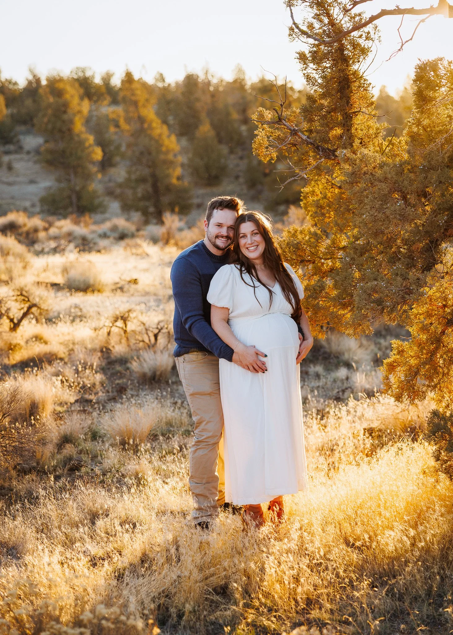 A couple stands together outdoors during sunset, smiling. The pregnant woman wears a white dress, and the man wears a navy sweater and beige pants. They are surrounded by tall grass and trees.