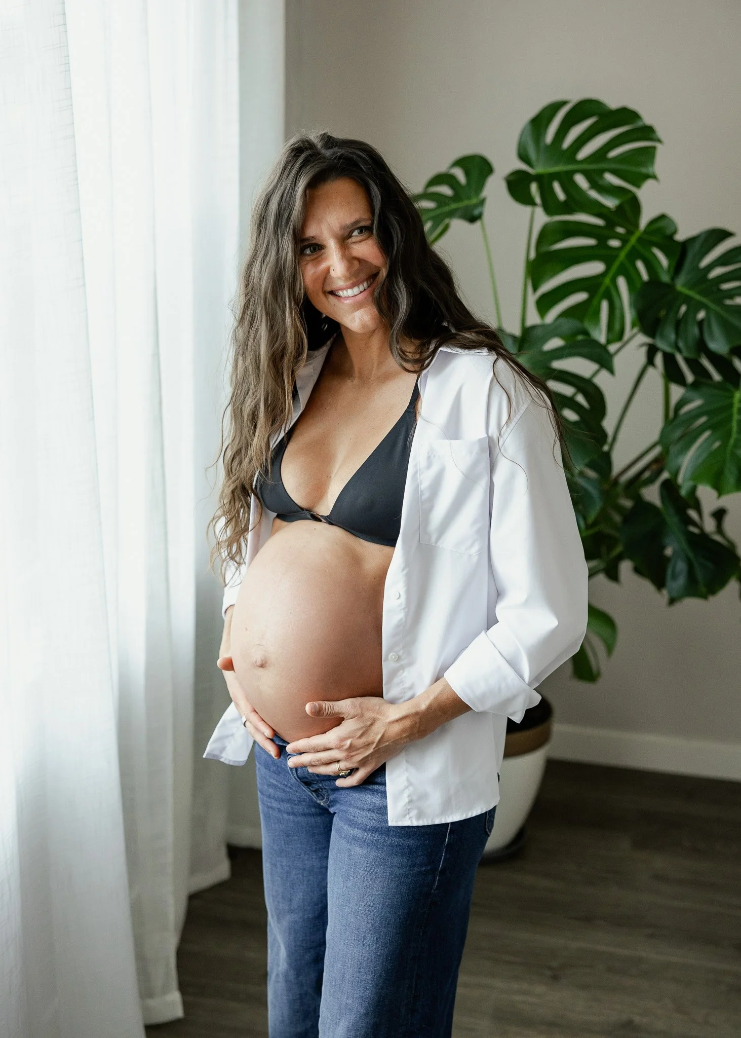 A pregnant woman smiling, wearing a black bra and a white shirt open, standing next to a potted plant in a well-lit room.