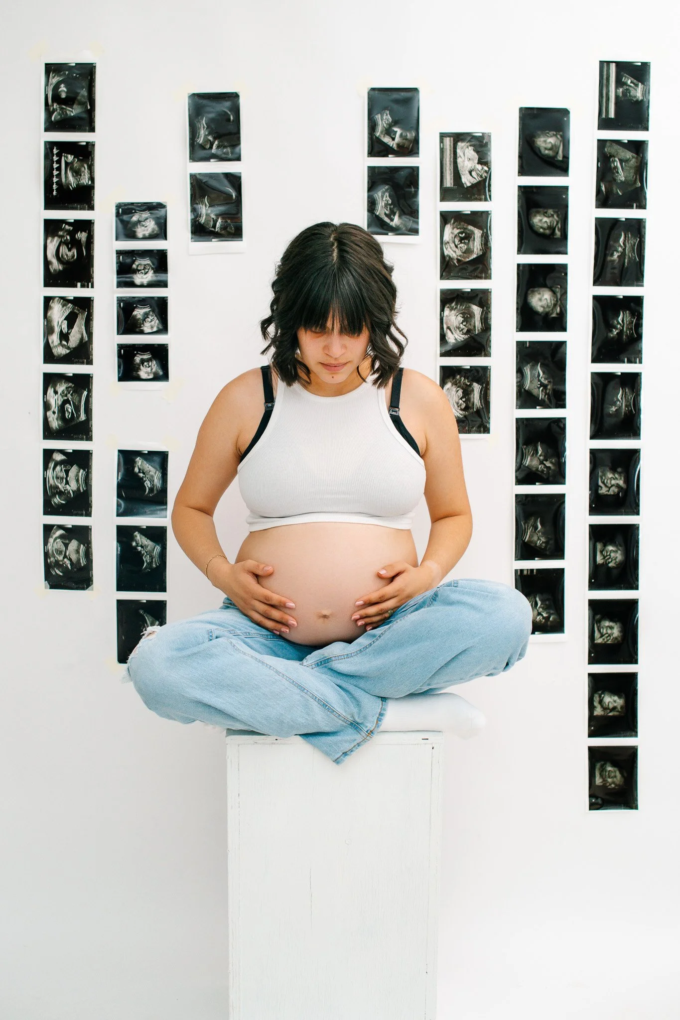 A pregnant woman sitting cross-legged on a white platform, holding her belly, with ultrasound images displayed on the wall behind her.
