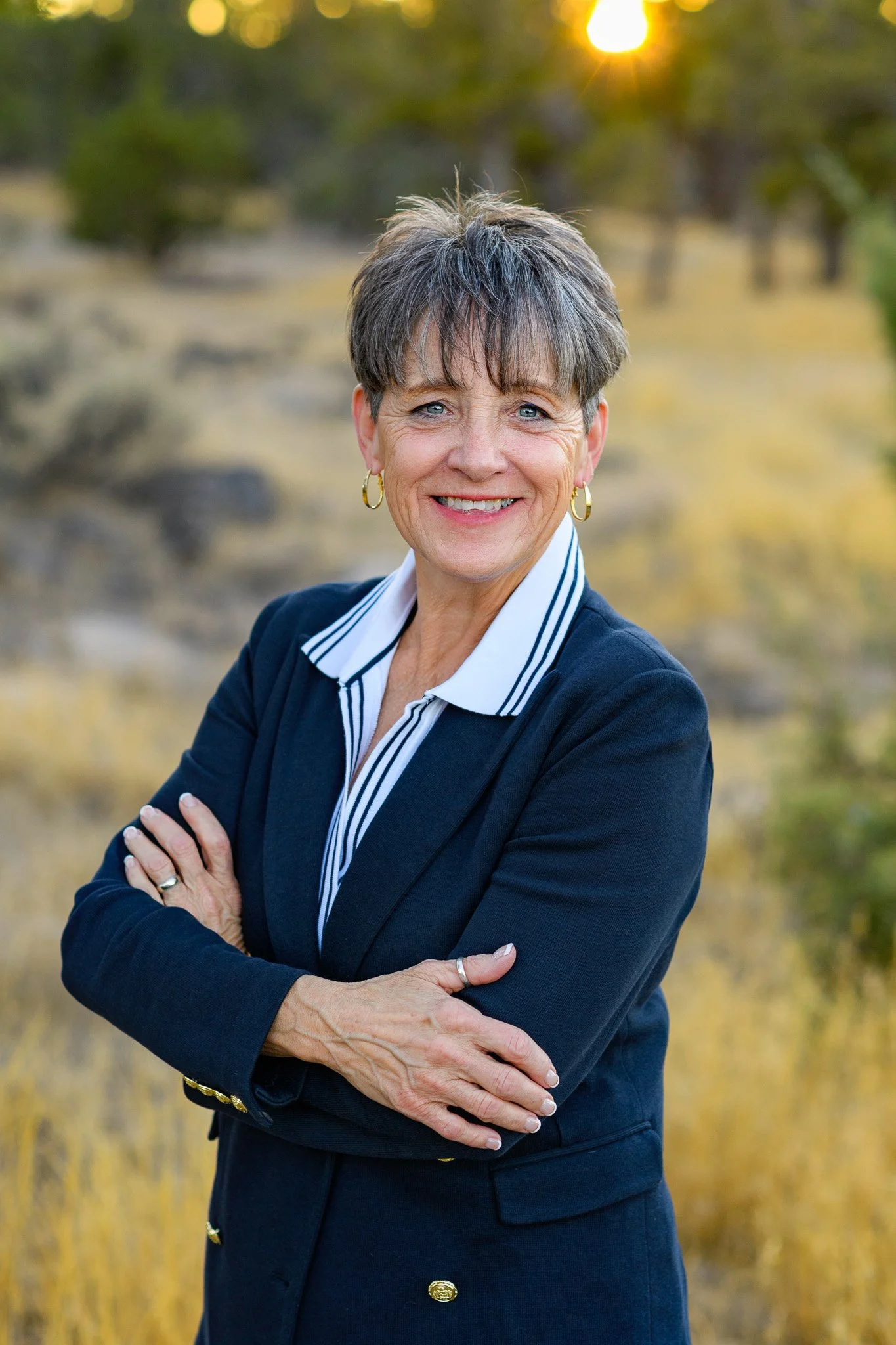 A smiling middle-aged woman in a navy blazer and striped shirt outdoors during sunset, with crossed arms, blonde and gray hair, and gold jewelry.