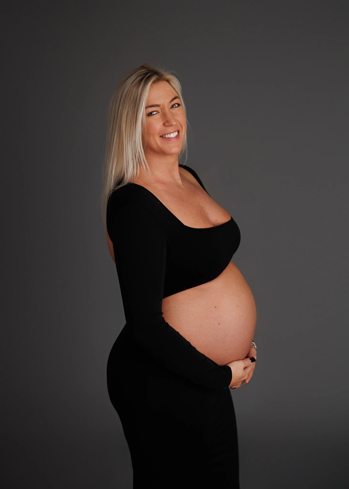 A pregnant woman with blonde hair smiling and wearing a black long-sleeve top and black skirt, holding her belly against a dark gray background.