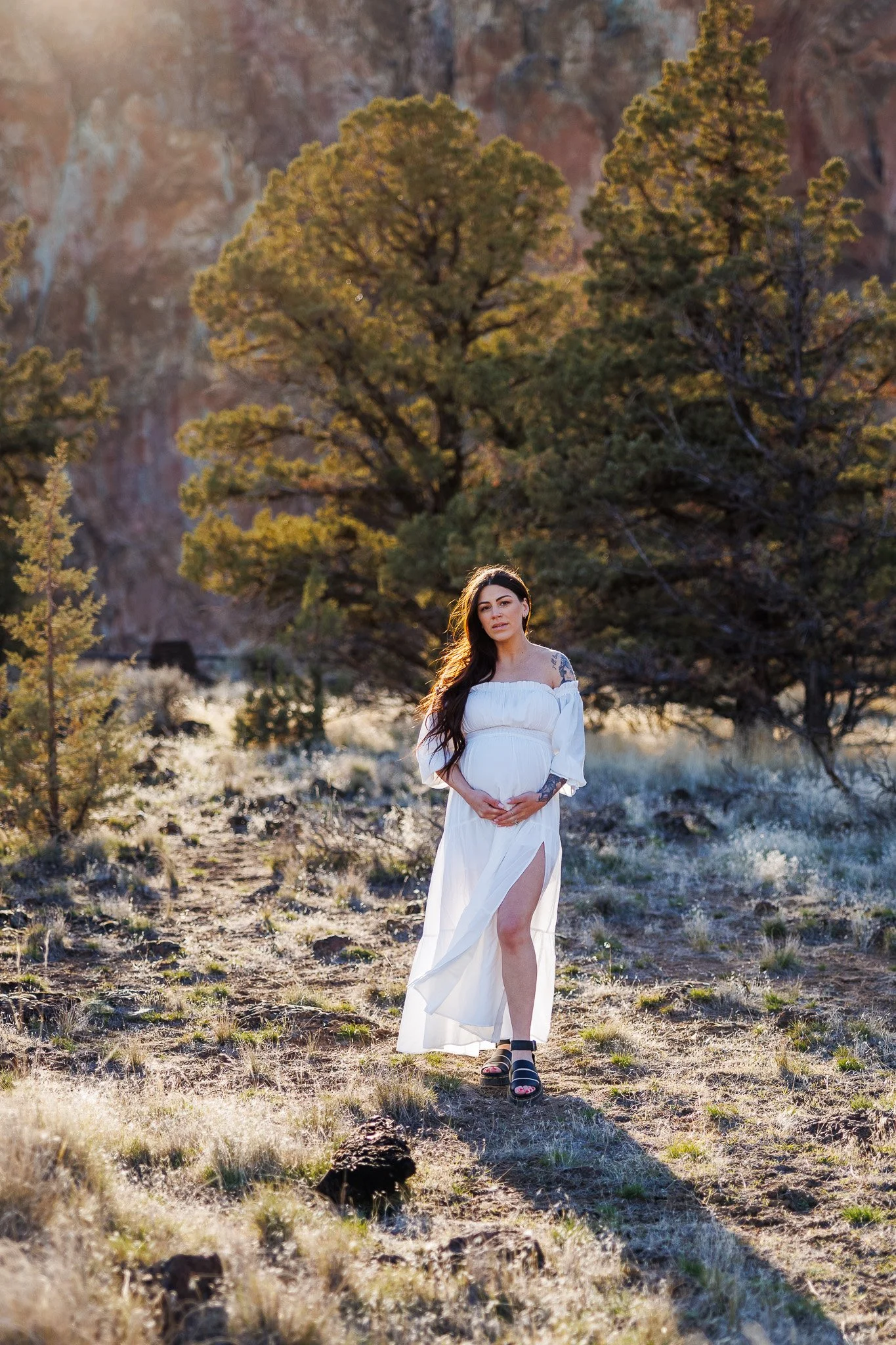 A pregnant woman in a white dress walking outdoors in a desert landscape with trees and rock formations in the background.