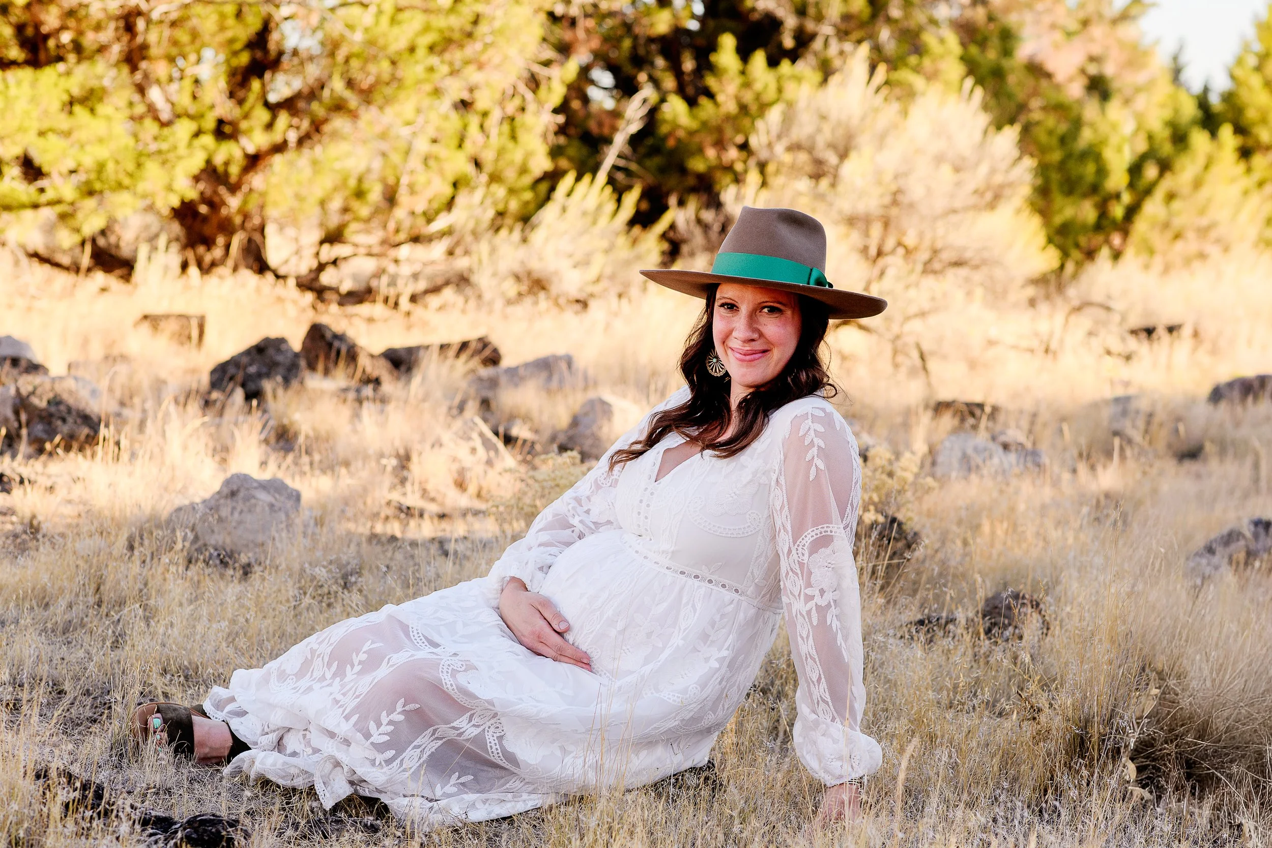 Pregnant woman in a white lace dress sitting on dry grass with rocks and trees in the background, wearing a wide-brimmed hat with a green band, smiling at the camera.