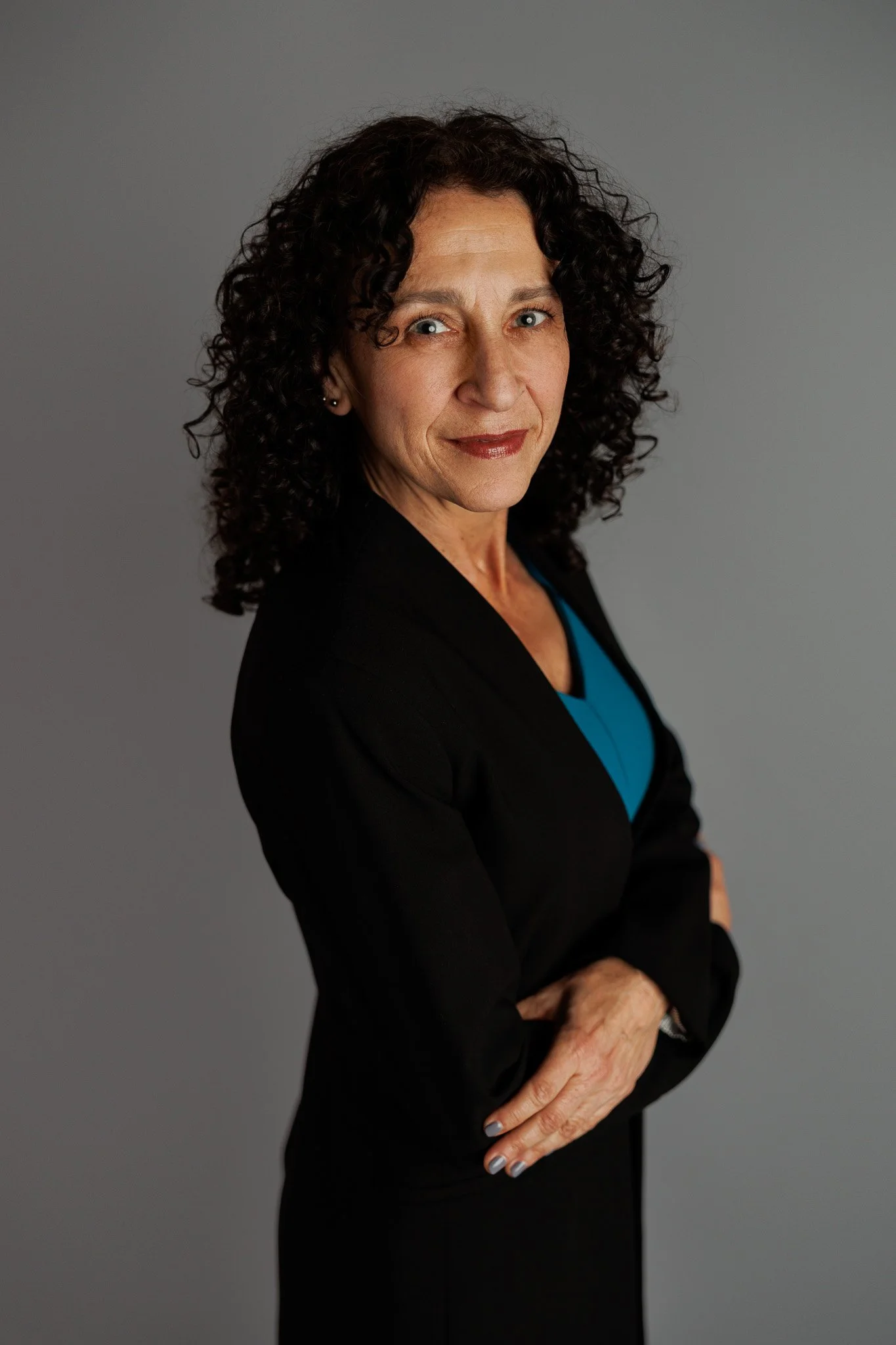 A portrait of a middle-aged woman with curly dark hair, wearing a black blazer over a blue top, standing with her arms crossed against a neutral gray background.