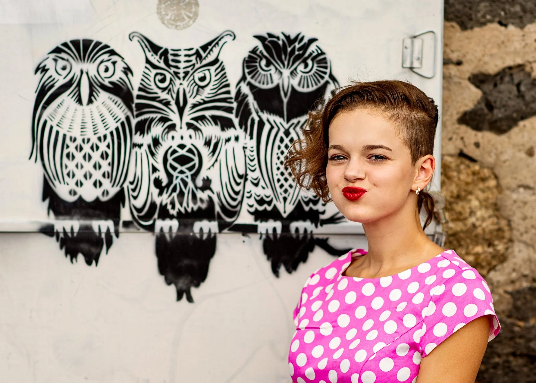 A woman with short curly hair wearing a pink dress with white polka dots, standing in front of a mural of black and white stylized owls and an owl with a dragonfly in the background.