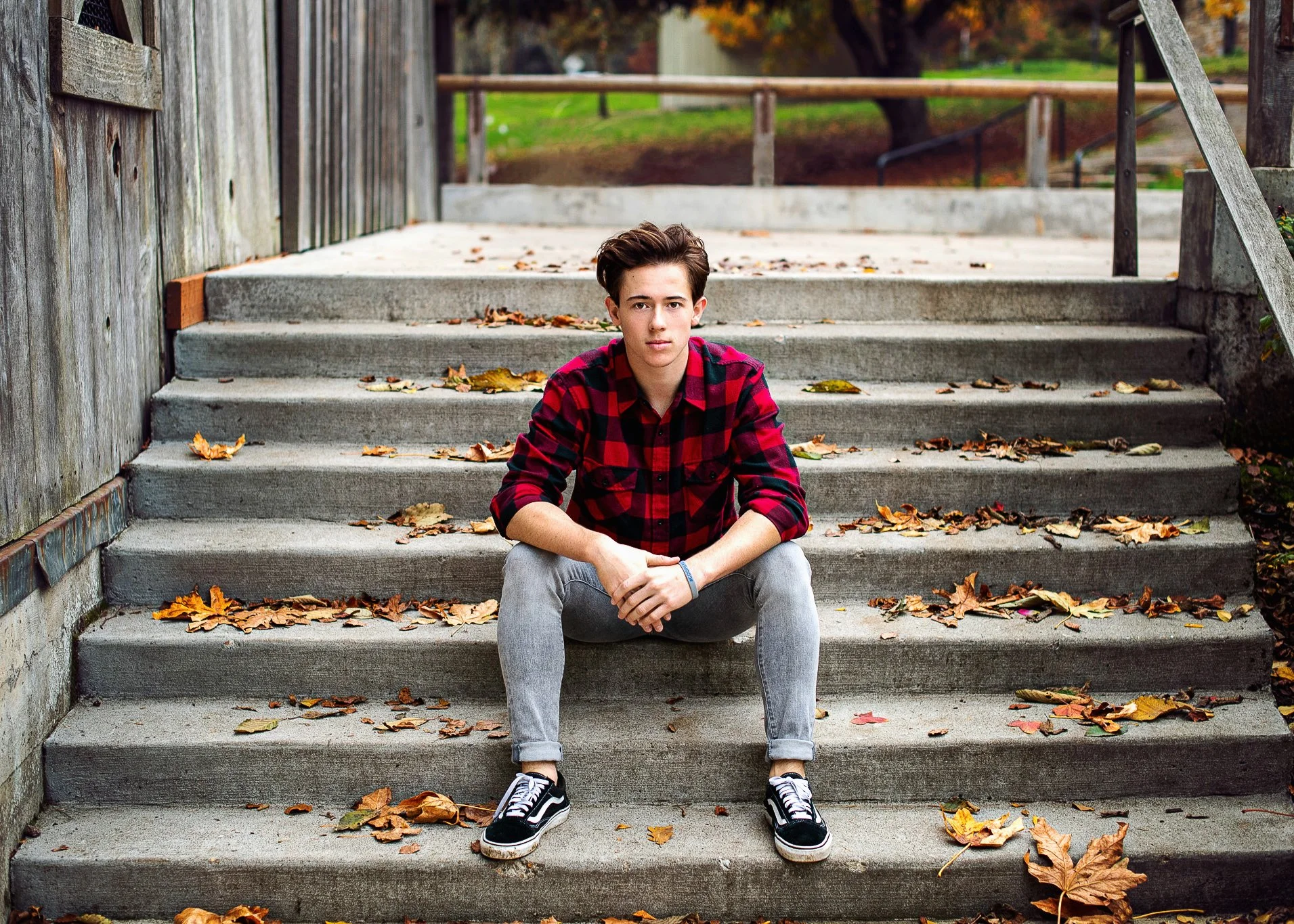 A young man with dark hair wearing a red and black plaid shirt and gray jeans is sitting on concrete stairs covered with fallen autumn leaves, outdoors with a park background.