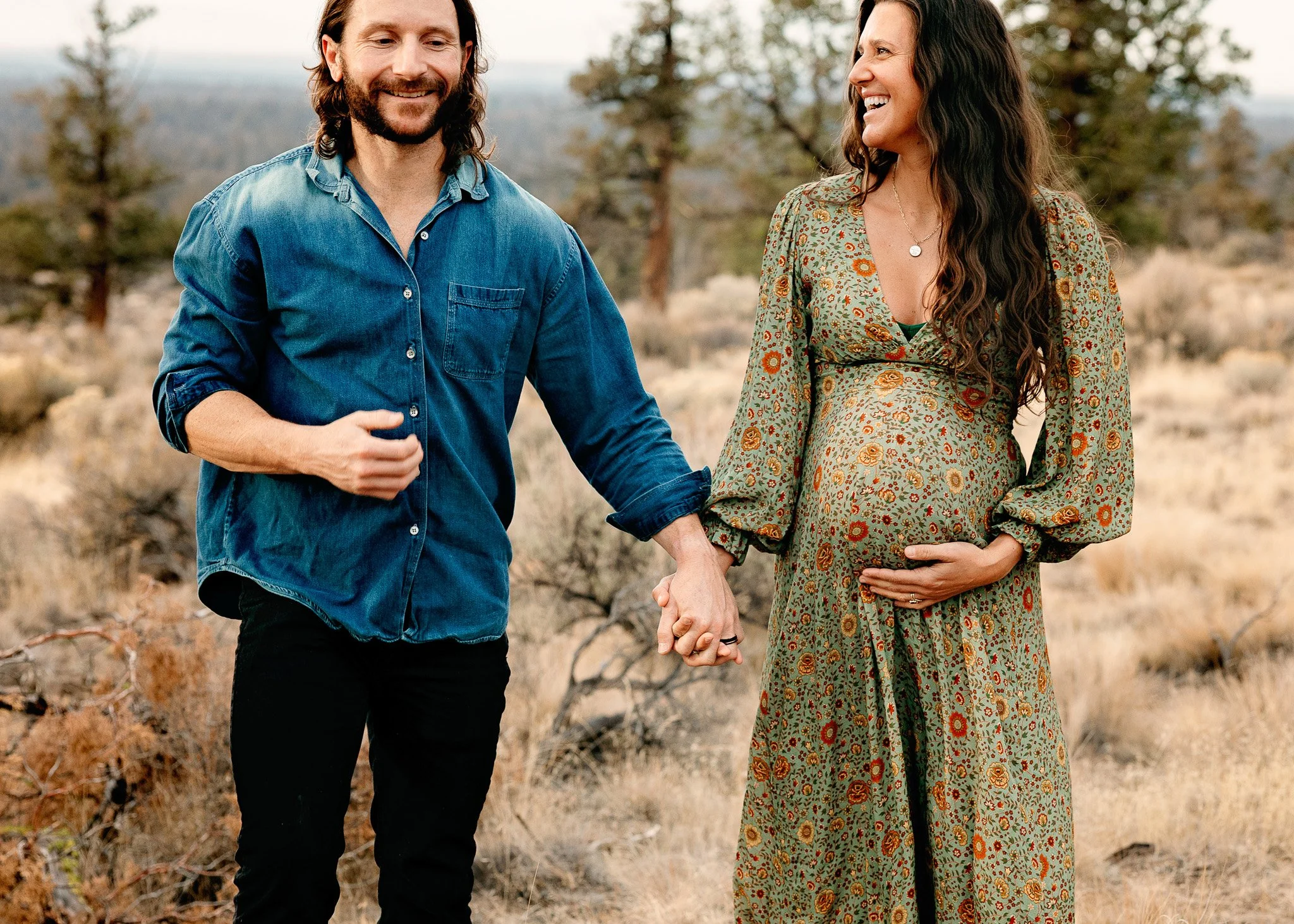 A smiling pregnant woman wearing a floral dress holding her baby bump with her right hand while holding her partner's hand with her left, walking in a desert landscape with trees in the background.