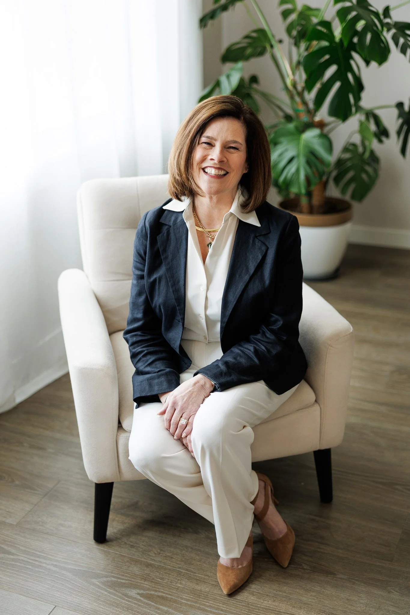 A woman with shoulder-length brown hair smiling, sitting on a cream-colored armchair in a bright room with a large green houseplant behind her.