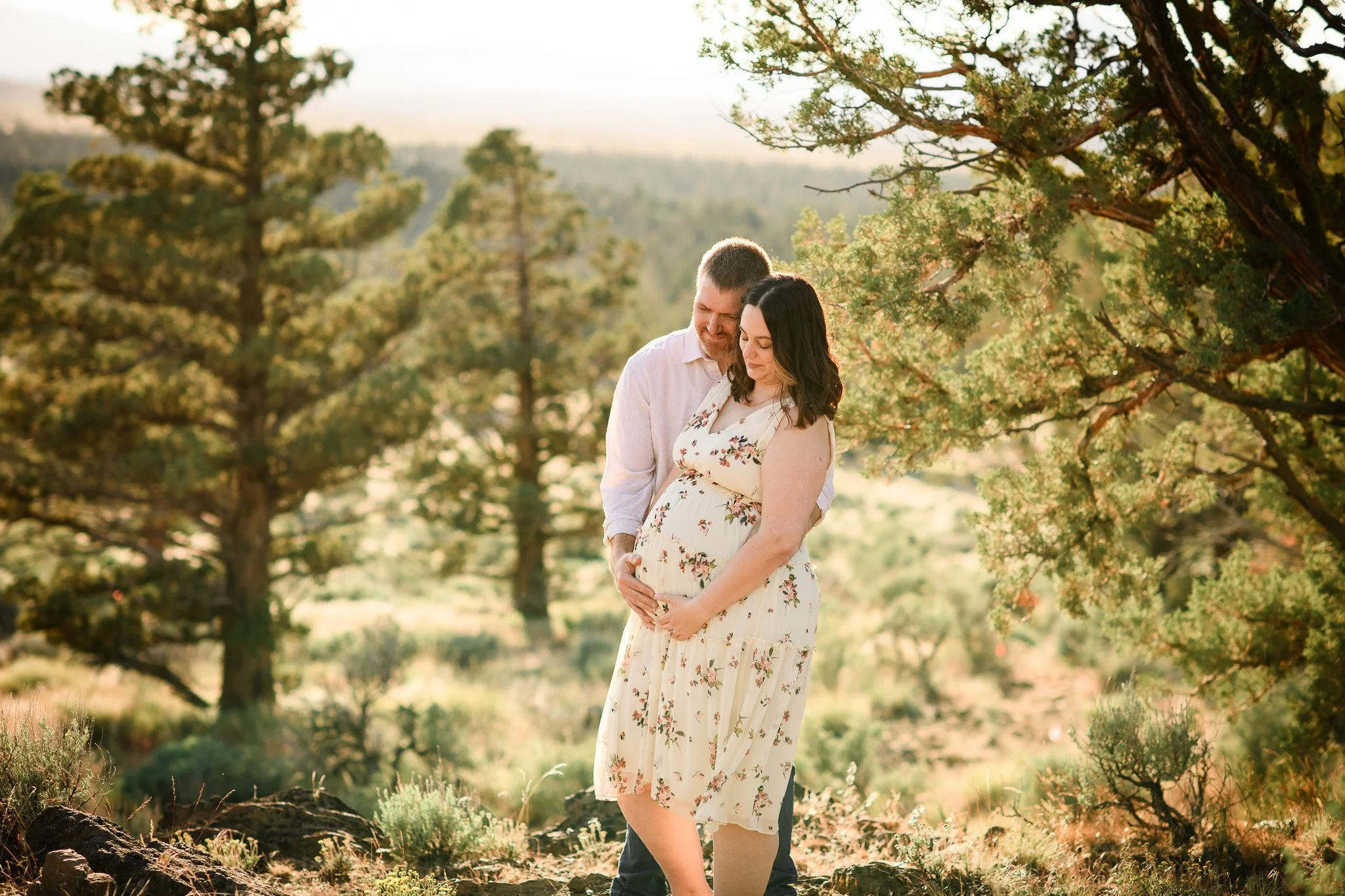 A couple, expecting a baby, standing in a sunny outdoor natural setting with trees and grass, lovingly holding the woman's pregnant belly.